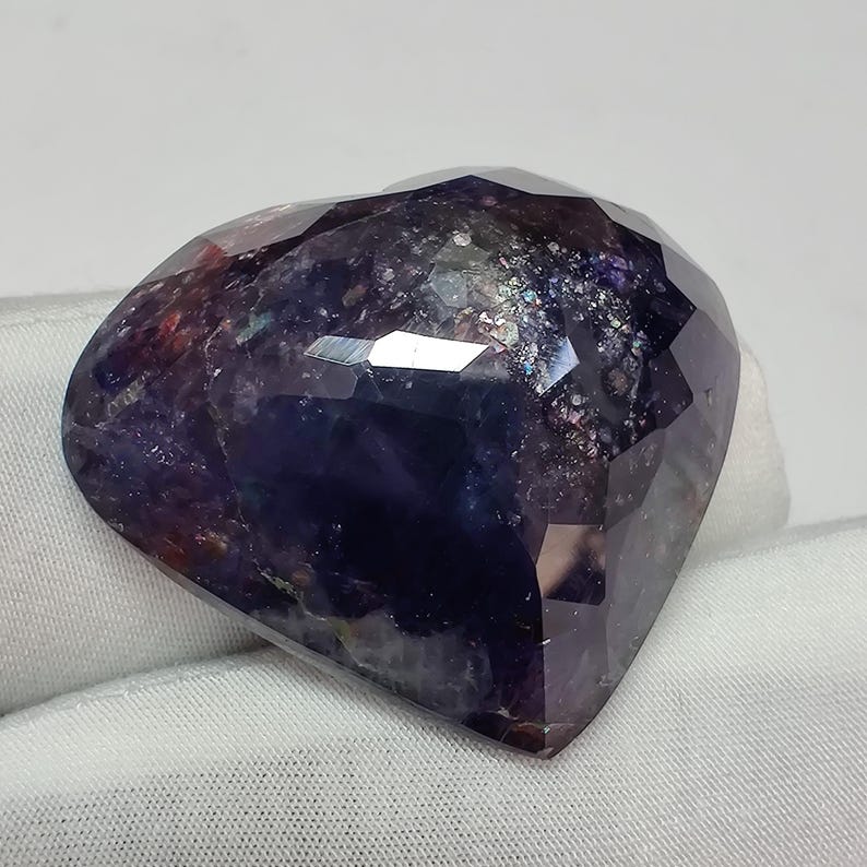 May include: A faceted, heart-shaped gemstone with a deep purple hue, exhibiting a play of colors. The stone's surface reflects light, showcasing a mix of dark blues, purples, and hints of red. The gemstone is set against a white background.