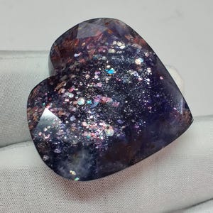 May include: A heart-shaped gemstone with a faceted surface. The stone has a deep purple base color with sparkling inclusions of various colors, including pink, blue, and silver. The gemstone is set against a white background.