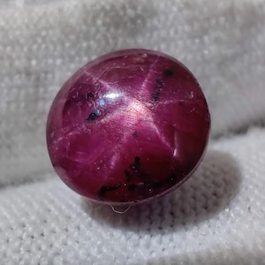 May include: A polished, cabochon-cut ruby gemstone. The deep red gem has a six-ray star effect, with dark inclusions. The stone is set against a white, textured background.