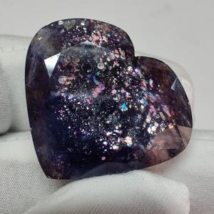 May include: A heart-shaped gemstone with a dark purple base color and iridescent inclusions of pink, blue, and silver. The faceted surface reflects light, creating a sparkling effect. The gemstone is held against a white fabric background.