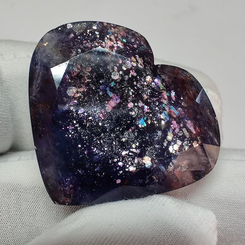 May include: A heart-shaped gemstone with a dark purple base color and iridescent inclusions of pink, blue, and silver. The faceted surface reflects light, creating a sparkling effect. The gemstone is held against a white fabric background.