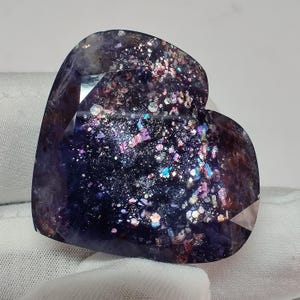 May include: A heart-shaped gemstone with a dark purple base color and iridescent inclusions. The stone has a faceted surface and displays a range of colors, including pink, blue, and silver. The gemstone is set against a white background.
