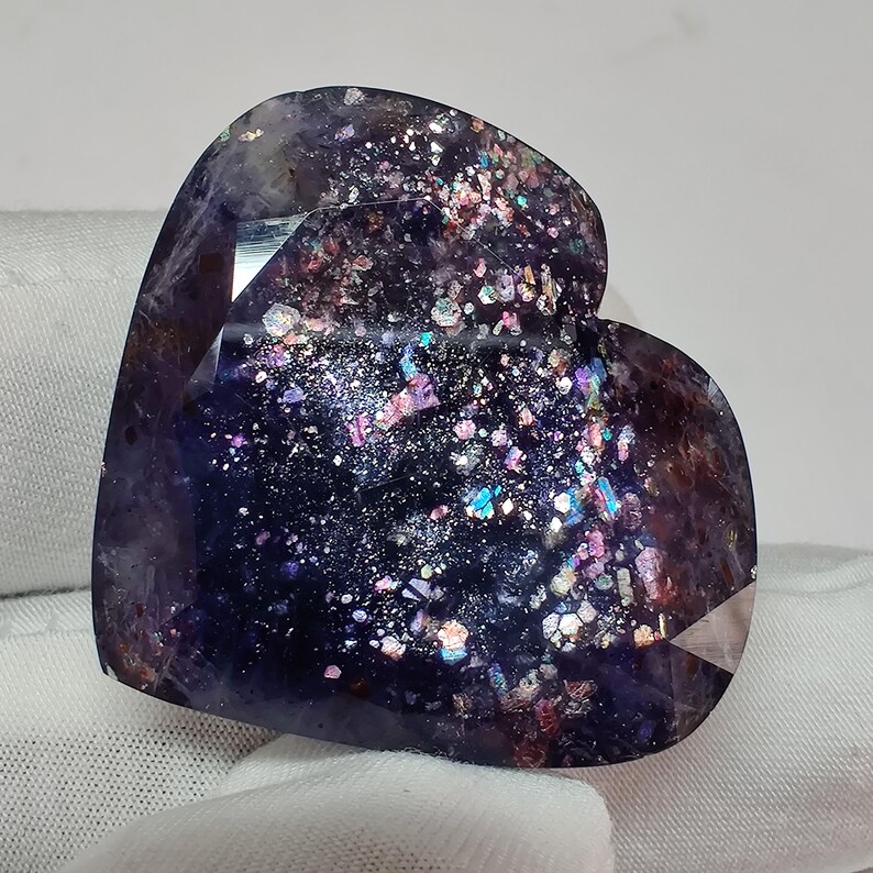 May include: A heart-shaped gemstone with a dark purple base color and iridescent inclusions. The stone has a faceted surface and displays a range of colors, including pink, blue, and silver. The gemstone is set against a white background.
