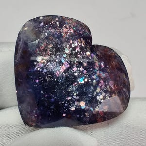 May include: A heart-shaped gemstone with a dark purple base color, speckled with iridescent glitter in shades of pink, blue, and silver. The stone has a faceted surface, catching light and creating a sparkling effect. The gemstone is displayed on a white surface.