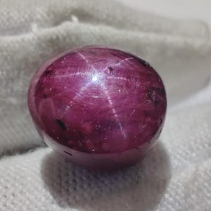 Può includere: Una gemma sferica lucidata, di un profondo colore magenta. La superficie mostra un effetto stella a sei raggi, con la luce che si riflette sulla pietra. La gemma è posta su uno sfondo bianco strutturato.