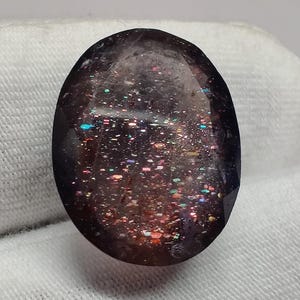 May include: A dark purple oval gemstone with iridescent inclusions. The gemstone has a speckled appearance with a variety of colors, including pink, red, and blue.