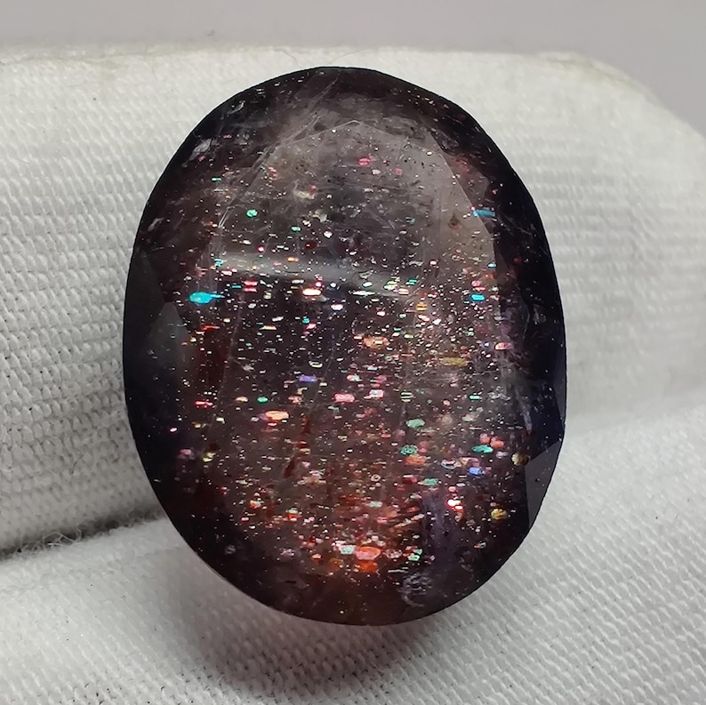 May include: A dark purple oval gemstone with iridescent inclusions. The gemstone has a speckled appearance with a variety of colors, including pink, red, and blue.
