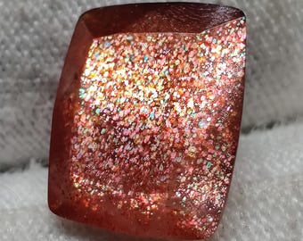 May include: A faceted, rectangular sunstone gemstone. The stone has a reddish-brown hue with sparkling inclusions of various colours, including pink, gold, and green. The gemstone is cut with slightly rounded corners.