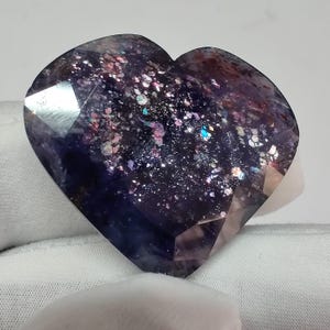 May include: A faceted, heart-shaped gemstone with a deep purple base color. The stone is speckled with iridescent flecks of pink, blue, and silver, creating a galaxy-like effect. The gem is held against a white background.