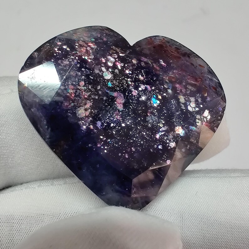 May include: A faceted, heart-shaped gemstone with a deep purple base color. The stone is speckled with iridescent flecks of pink, blue, and silver, creating a galaxy-like effect. The gem is held against a white background.