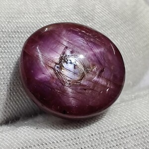 Peut inclure: Une pierre précieuse de rubis polie, de forme ovale. La gemme est d'un rouge-violet profond avec un effet étoilé visible sur sa surface. La pierre a une surface lisse et réfléchissante avec des inclusions internes.