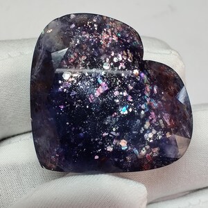 May include: A heart-shaped gemstone with a dark blue base and sparkling inclusions of various colors. The polished surface reflects light, creating a galaxy-like effect. The gemstone is approximately 2 inches wide.