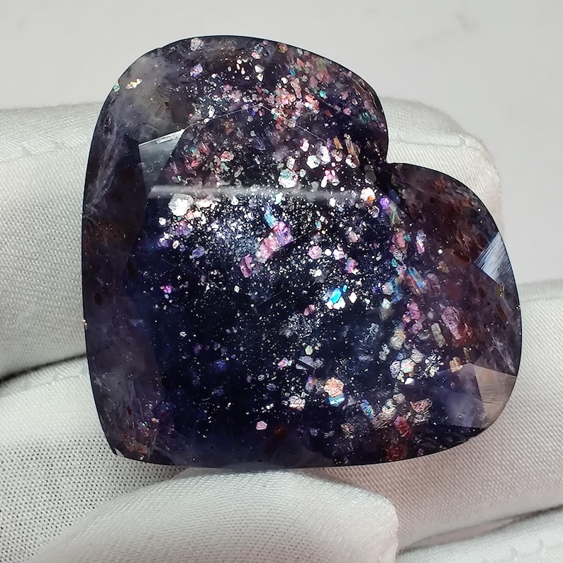 May include: A heart-shaped gemstone with a dark blue base and sparkling inclusions of various colors. The polished surface reflects light, creating a galaxy-like effect. The gemstone is approximately 2 inches wide.