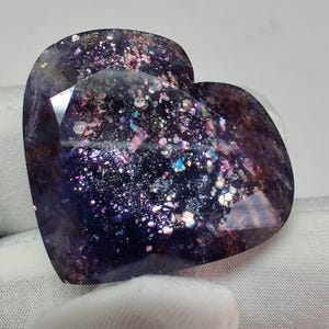 May include: A heart-shaped gemstone with a faceted surface. The stone is primarily dark purple with iridescent flecks of pink, blue, and gold. The gemstone is held against a white background.