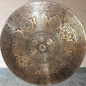 May include: A large, circular brass gong featuring intricate laser-etched designs. The center displays a seated figure surrounded by chakra symbols and a Greek key border. Concentric rings contain geometric patterns, flower of life motifs, and caduceus symbols.