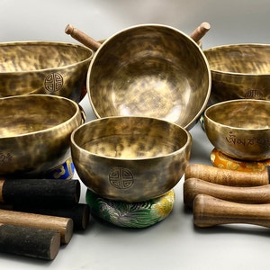 May include: A set of seven singing bowls with various sizes and designs. The bowls are made of metal and have a hammered finish. Each bowl has a unique inscription and comes with a wooden striker. The bowls are arranged on a white surface with cushions.