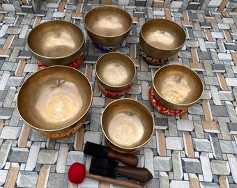 May include: Set of seven singing bowls with colorful cushions and two wooden mallets with red felt tips. The bowls are made of metal and have a shiny finish.