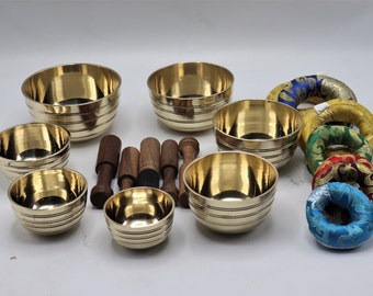 May include: A set of seven brass singing bowls with wooden strikers and colorful fabric cushions. The bowls are of varying sizes and have a hammered finish.