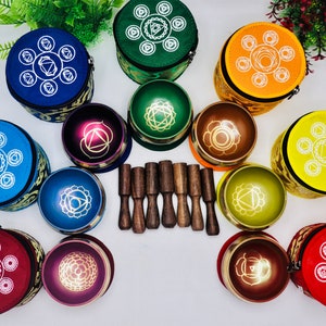 Op de afbeelding: Zeven chakra-zingende kommen met hamers, elke kom heeft een andere kleur en een uniek ontwerp. De kommen zijn in een cirkel gerangschikt met de hamers in het midden. De kommen zijn gemaakt van metaal en hebben een glanzende afwerking.