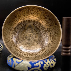 May include: A golden singing bowl with intricate engravings and a depiction of a deity in the center. The bowl is resting on a blue and gold cushion.