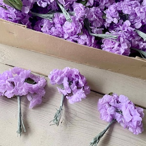 May include: A box of lavender silk flower bunches. The bunches are made of soft, delicate fabric and have a realistic appearance. They are perfect for adding a touch of elegance to any craft project or floral arrangement.