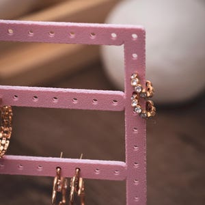 Puede incluir: Un expositor de pendientes rosa con varios pares de pendientes dorados. Un par tiene piedras transparentes, otro es un aro de estilo cadena. El expositor est&aacute; hecho de pl&aacute;stico rosa.