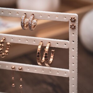 Puede incluir: Exhibici&oacute;n de varios pendientes en un soporte blanco. Los pendientes incluyen peque&ntilde;os pendientes de oro, aros dorados con detalles cuadrados y aros dorados con piedras transparentes. Algunos pendientes tienen un acento de perla.