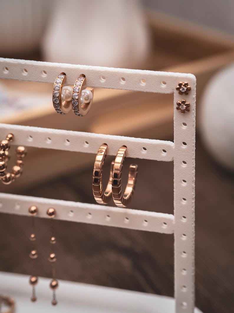 Puede incluir: Exhibici&oacute;n de varios pendientes en un soporte blanco. Los pendientes incluyen peque&ntilde;os pendientes de oro, aros dorados con detalles cuadrados y aros dorados con piedras transparentes. Algunos pendientes tienen un acento de perla.