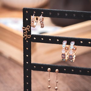 Puede incluir: Un expositor de pendientes negro que muestra varios pendientes. Los pendientes incluyen peque&ntilde;os pendientes de lazo dorado, pendientes de aro con piedras transparentes y pendientes de aro con gemas de colores. Tambi&eacute;n hay pendientes colgantes largos y delicados con peque&ntilde;as cuentas doradas.