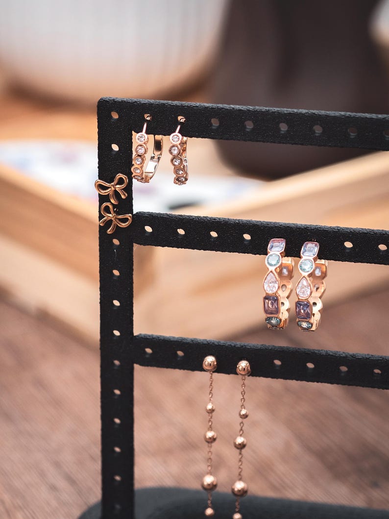 Puede incluir: Un expositor de pendientes negro que muestra varios pendientes. Los pendientes incluyen peque&ntilde;os pendientes de lazo dorado, pendientes de aro con piedras transparentes y pendientes de aro con gemas de colores. Tambi&eacute;n hay pendientes colgantes largos y delicados con peque&ntilde;as cuentas doradas.