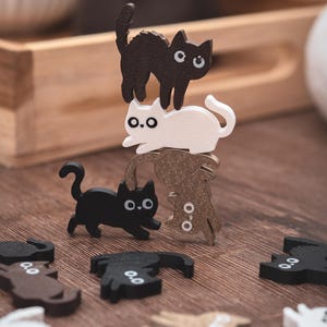 Op de afbeelding: Houten kattenbeeldjes in zwart, wit en bruin, gestapeld en verspreid op een houten oppervlak. De katten hebben eenvoudige vormen en grote, ronde ogen. De opstelling creëert een speelse, evenwichtige compositie.