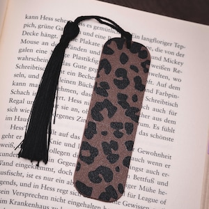 Puede incluir: Un marcapáginas con estampado de leopardo marrón y negro con una borla negra. El marcapáginas es rectangular y está hecho de un material similar al cuero. Se coloca encima de un libro abierto.