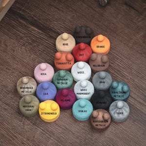 Puede incluir: Una colecci&oacute;n de peque&ntilde;os objetos redondos y coloridos con una superficie texturizada y un peque&ntilde;o bot&oacute;n en la parte superior. Cada objeto est&aacute; etiquetado con un nombre de color diferente, incluyendo "Naranja", "Azul" y "Verde". Los objetos est&aacute;n dispuestos sobre una superficie de madera.