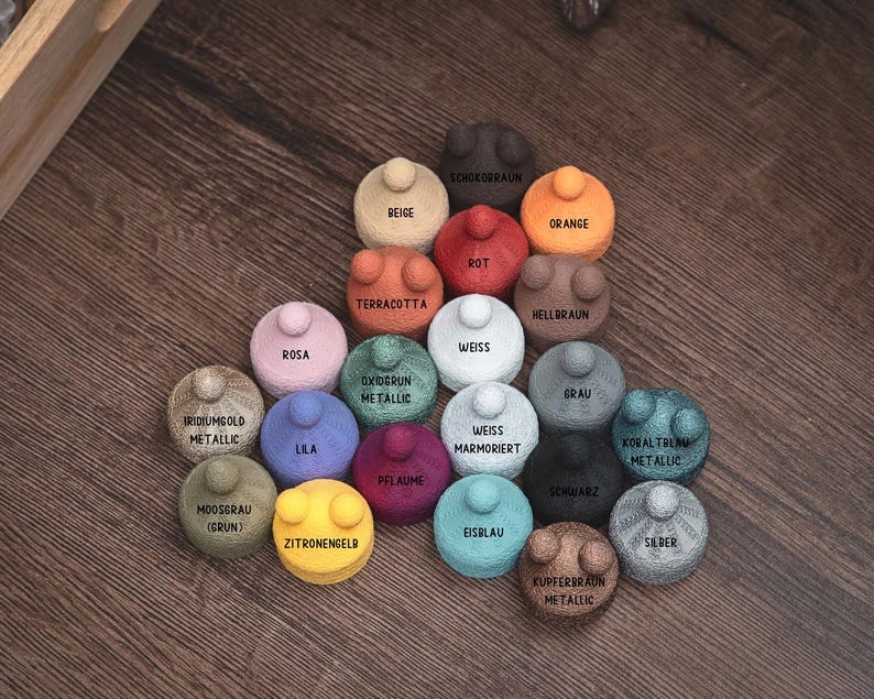 Puede incluir: Una colecci&oacute;n de peque&ntilde;os objetos redondos y coloridos con una superficie texturizada y un peque&ntilde;o bot&oacute;n en la parte superior. Cada objeto est&aacute; etiquetado con un nombre de color diferente, incluyendo "Naranja", "Azul" y "Verde". Los objetos est&aacute;n dispuestos sobre una superficie de madera.