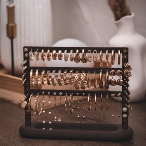 Puede incluir: Un expositor de pendientes marr&oacute;n oscuro lleno de varios pendientes dorados. Los pendientes incluyen aros, colgantes y tachuelas, mostrando una variedad de dise&ntilde;os y adornos. El expositor est&aacute; sobre una superficie de madera.