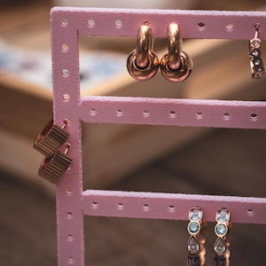 Puede incluir: Un expositor de pendientes rosa con varios pares de pendientes dorados. Los pendientes incluyen aros, dise&ntilde;os geom&eacute;tricos y adornos de piedras preciosas. El expositor es de pl&aacute;stico rosa y tiene varias filas de agujeros para exhibir pendientes.