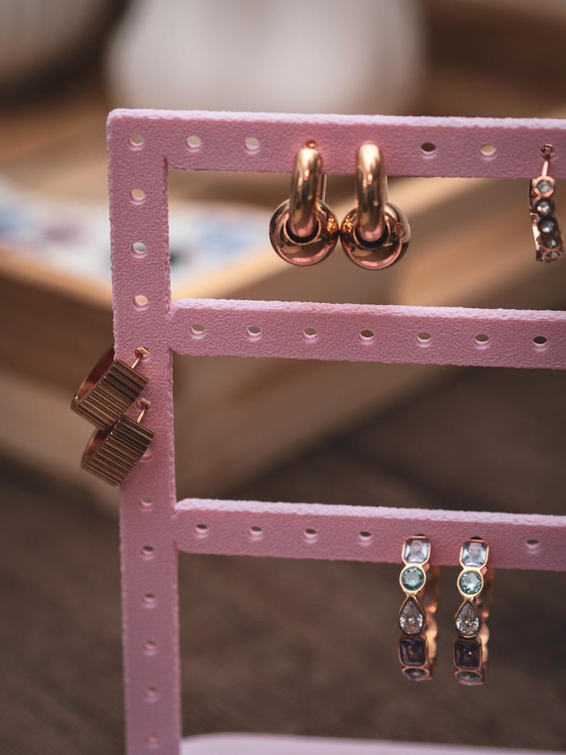 Puede incluir: Un expositor de pendientes rosa con varios pares de pendientes dorados. Los pendientes incluyen aros, dise&ntilde;os geom&eacute;tricos y adornos de piedras preciosas. El expositor es de pl&aacute;stico rosa y tiene varias filas de agujeros para exhibir pendientes.