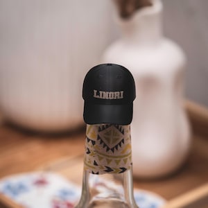 Op de afbeelding: Een heldere glazen fles met een patroonlabel en een zwarte dop met het woord "LINORI". De fles staat op een houten dienblad. De achtergrond is wazig.