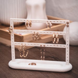 Puede incluir: Un expositor de pendientes blanco con varios pendientes dorados. Los pendientes incluyen aros, tachuelas y modelos colgantes. El expositor est&aacute; sobre una superficie de madera, con un fondo borroso.