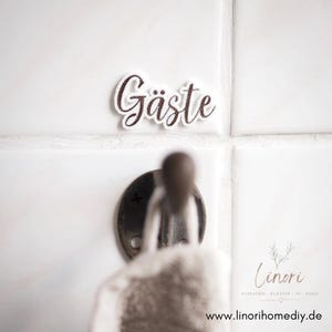 Könnte beinhalten: Weiße Fliesenwand mit einem dekorativen Schild mit der Aufschrift "Gäste" in brauner Schrift. Darunter befindet sich ein Metallhaken, an dem ein Handtuch hängt. Das Bild zeigt auch ein Logo und eine Website-Adresse.