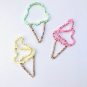 May include: Three knitted ice cream cone decorations in pastel shades. The ice cream scoops are light green, pink, and yellow, with brown cone bases. They are arranged on a white background.
