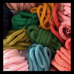 May include: A close-up of a collection of knitted cords in a variety of colours, including pink, green, yellow, orange, and blue. The cords are arranged together, highlighting their texture and vibrant hues. The cords appear to be made of a thick yarn.