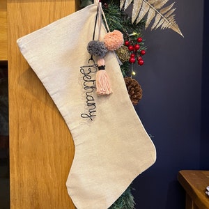 May include: A beige Christmas stocking with the name "Bethany" embroidered in black. The stocking is decorated with pink and grey pom-poms and pink tassels.