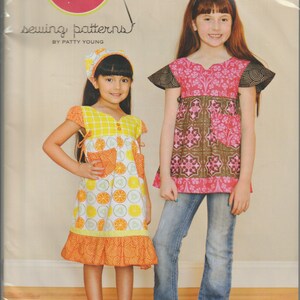 May include: A sewing pattern package showing two girls in tunic dresses and shirts with headscarves. The package includes the text "Frida" and "modkid", and the sizes are 2T-10. The dresses have bright, colourful patterns.