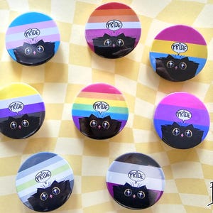 May include: A selection of Pride-themed badges. Each badge showcases a black cat illustration and the word "PRIDE" in a speech bubble. The badges feature different Pride flag designs, including rainbow, transgender, and non-binary flags.
