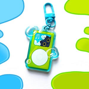 May include: A green and white keychain shaped like a music player, featuring a cartoon cat with yellow eyes. The keychain has a blue clasp and is surrounded by bubble-like shapes. The background has blue and green abstract designs.