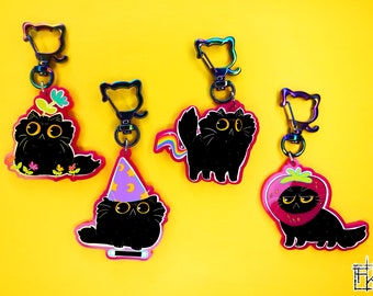 Könnte beinhalten: Satz mit fünf schwarzen Katzen-Schlüsselanhängern mit verschiedenen Designs. Jeder Schlüsselanhänger zeigt eine schwarze Katze mit einem anderen Accessoire, darunter eine Blumenkrone, einen Hexenhut, einen Regenbogen-Schwanz, einen Erdbeerhut und ein schlichtes Design. Die Schlüsselanhänger sind aus Metall und haben eine regenbogenfarbene, irisierende Oberfläche.
