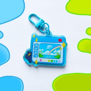 May include: A blue keychain featuring a cartoon camera design with a beach scene and a rainbow. The keychain has a matching blue clasp and a small cat peeking out from behind the camera. The design includes yellow star accents.
