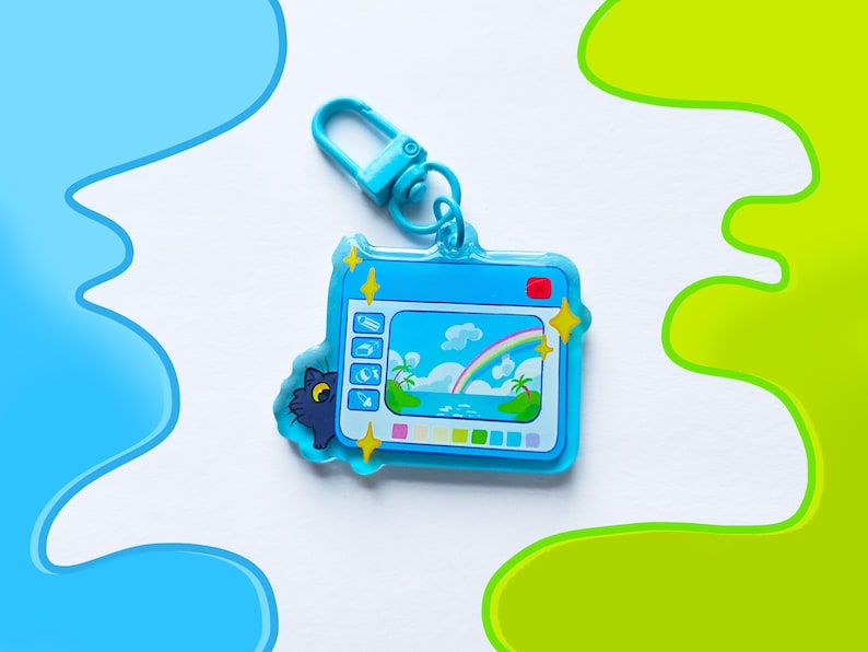 May include: A blue keychain featuring a cartoon camera design with a beach scene and a rainbow. The keychain has a matching blue clasp and a small cat peeking out from behind the camera. The design includes yellow star accents.