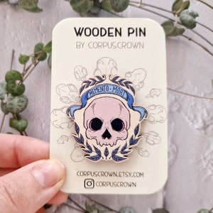 May include: Wooden pin featuring a skull with a blue banner that reads "Memento Mori" and a wreath of leaves. The pin is on a white card with the text "Wooden Pin by Corpuscrown" and the website address "corpuscrown.etsy.com".